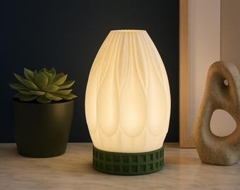 Puede incluir: Una lámpara de mesa blanca e iluminada con un diseño texturizado en forma de pétalo. La lámpara tiene una base verde geométrica y se encuentra sobre una superficie blanca. También se ven una planta suculenta en maceta y una escultura de piedra.
