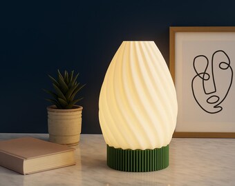 Puede incluir: Lámpara en forma de huevo con diseño en espiral crema y base verde. En el fondo se ven un cuadro con un dibujo lineal y una planta suculenta en maceta, junto a un libro cerrado.