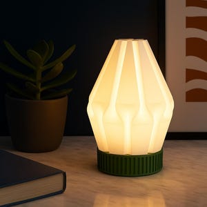 May include: A modern, illuminated table lamp with a white, geometric shade and a green base. The lamp is turned on, emitting a warm glow. A dark blue book and a potted plant are in the background.