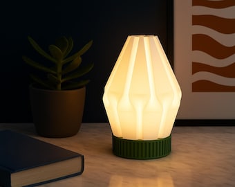 Puede incluir: Una lámpara de mesa moderna con una pantalla blanca geométrica y una base verde. La lámpara está encendida, emitiendo un brillo cálido. Un libro azul oscuro y una planta en maceta están en el fondo.