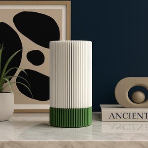 May include: A modern interior design scene featuring a white and green cylindrical object with vertical lines, a potted plant, and a framed abstract art piece. A beige sculpture sits atop a book that reads "ANCIENT ART".