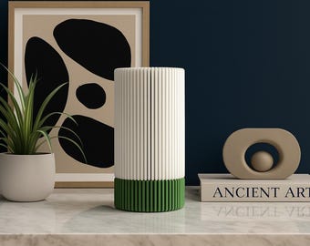 Puede incluir: Una escena de diseño de interiores moderno con un objeto cilíndrico blanco y verde con líneas verticales, una planta en maceta y una obra de arte abstracta enmarcada. Una escultura beige se encuentra sobre un libro con "ANCIENT ART".
