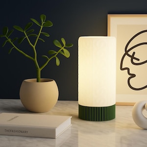 May include: A lit cylindrical lamp with a green base sits on a marble surface, next to a potted plant and a book. A framed abstract face art piece and a white sculpture are also present. The lamp emits a soft glow.
