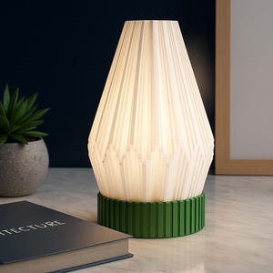 May include: A modern table lamp with a white, textured shade and a green base. The lamp is turned on, emitting a soft glow. A succulent plant in a gray pot and a book titled "ARCHITECTURE" are also visible.
