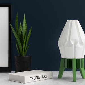 May include: A white geometric lamp with green accents, a black framed picture, a potted snake plant, a white book with the word "TREESSENCE", and a white decorative sculpture. The lamp has a unique design and is on a white surface.