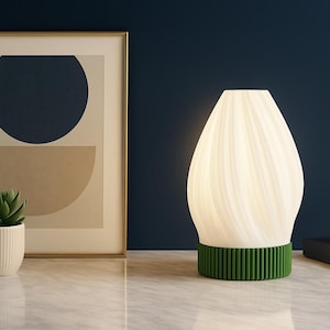 May include: A modern white lamp with a green base sits on a marble surface. A framed abstract art print and a small succulent in a white pot are to the left. A white sculpture and a book are to the right.