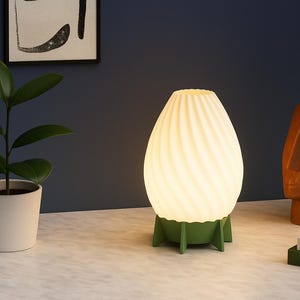 May include: A glowing white and green table lamp with a spiral design. The lamp is egg-shaped and sits on a green base. A small potted plant and a brown sculpture are also visible.