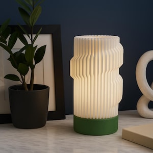 May include: A modern white table lamp with a green base, featuring a ribbed design. A potted plant in a black pot and a framed picture are in the background. A white sculpture and a beige book are also visible.