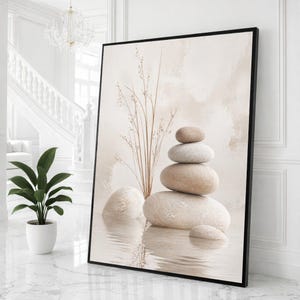 Op de afbeelding: Een ingelijste kunstdruk met een zen-geïnspireerde compositie. Het toont een stapel gladde, beige stenen in water, met lange, slanke grassen en een zachte, neutrale achtergrond. Het kunstwerk is ingesloten in een zwart frame, wat een rustgevende en minimalistische esthetiek creëert.