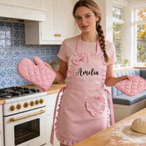 Pode incluir: Avental rosa com babados, um la&ccedil;o e o nome "Amelia" em escrita cursiva. Luva de forno e pegador de panela rosa combinando. O avental tem um bolso em forma de cora&ccedil;&atilde;o. Em uma cozinha.