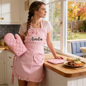 Pode incluir: Um avental rosa com babados e um la&ccedil;o, combinado com uma luva de forno e um descanso de panela a condizer. O avental &eacute; personalizado com o nome "Amelia". A cozinha inclui uma bancada de madeira e uma janela.