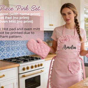 Pode incluir: Um conjunto de cozinha rosa de 3 pe&ccedil;as, incluindo um avental com o nome "Amelia", um pegador e uma luva de forno. O avental tem um la&ccedil;o e um bolso em forma de cora&ccedil;&atilde;o. O pegador e a luva t&ecirc;m um padr&atilde;o acolchoado.