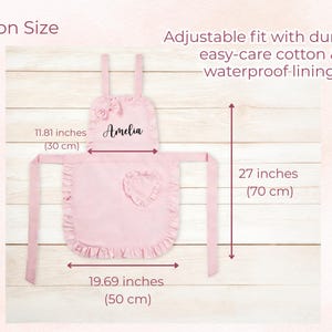 Pode incluir: Um avental rosa com borda franzida, la&ccedil;o e bolso em forma de cora&ccedil;&atilde;o. O avental tem 50 cm de largura, 70 cm de comprimento e uma largura superior de 30 cm. O avental tem o nome "Amelia" impresso.