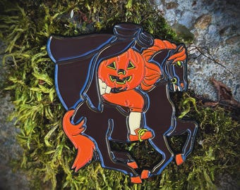 May include: Enamel pin featuring a pumpkin-headed figure riding a black horse. The figure is wearing orange clothing and a black cloak. The horse has orange hair and a blue outline. The pin is set against a mossy background.