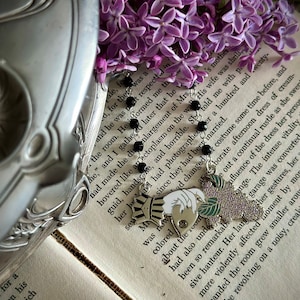 Puede incluir: Un brazalete de plata con cuentas negras y tres dijes. Los dijes son una mano sosteniendo un ramo de flores moradas, una mano sosteniendo un libro y una mano sosteniendo una corona. El brazalete está sobre un libro con texto sobre la madre de una mujer.