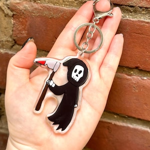 May include: A clear acrylic keychain featuring a cartoon Grim Reaper holding a scythe. The Reaper is black with a white skull and red eyes. The keychain has a silver metal ring and chain.