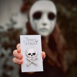 May include: White enamel pin featuring a skull and crossbones with the text "The Spawn of Verona" on a white card.
