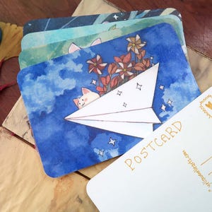 May include: A collection of illustrated postcards with watercolour designs. One card depicts a cat in a paper aeroplane against a blue sky. The word "POSTCARD" is visible on a blank card. A calligraphy pen and ink bottle are also present.