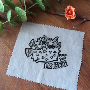 Peut inclure: Un carré de tissu gris avec une illustration en noir et blanc d'un poisson-globe et le texte "Keep your distance".