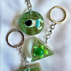 May include: Three keyrings with silver rings and chains. Each keyring features a clear resin pendant with a miniature figurine inside. The pendants include a dog, a turtle, and a mermaid. The dog is wearing blue swim shorts.