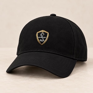 Può includere: Un cappellino da baseball nero con un emblema centrato sul pannello frontale. L'emblema è uno scudo con bordo dorato su sfondo nero, che mostra una grafica a cobra argentata e il testo "SHELBY" in lettere maiuscole bianche al centro.