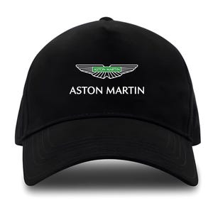 May include: Black baseball cap with the Aston Martin logo in white and green. The logo is a wing design, and the brand name is printed below in white capital letters. The cap has a curved peak.