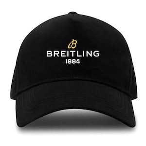 Könnte beinhalten: Schwarze Baseballkappe mit dem Breitling-Logo in Weiß und Gold. Das Logo enthält den Markennamen und das Jahr 1884. Die Kappe hat einen gebogenen Schirm und eine strukturierte Krone.