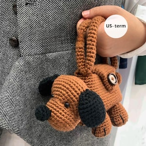 May include: A brown and black crocheted dog-shaped bag with button details. The dog has black ears and a black nose. The bag is held by a person wearing a gray herringbone vest.