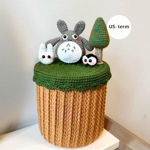 Op de afbeelding: Een handgemaakte gehaakte opbergcontainer met een grijze Totoro-personage, een wit konijn, een klein zwart wezen en een groene boom bovenop. De container heeft een groene bovenkant en een bruin, getextureerd lichaam. De tekst "US-term" staat in een witte cirkel.