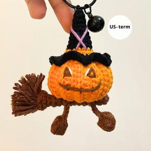 Op de afbeelding: Een handgemaakt gehaakt Halloween pompoen karakter met een heksenhoed, bezem en bel bedel. De oranje pompoen heeft een lachend gezicht en draagt een zwarte hoed met een roze kruis. De bezem en voeten zijn bruin.