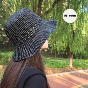 Op de afbeelding: Een zwarte geweven zonnehoed met een licht schuine rand. De hoed heeft een textuurpatroon en decoratieve accenten op de kroon. De hoed wordt gedragen door een persoon met lang bruin haar. De tekst "US-term" staat in een witte cirkel.