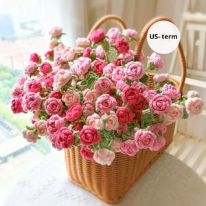 May include: A wicker basket filled with crocheted roses in various shades of pink and red. The roses are accompanied by green leaves, forming a colourful floral arrangement. The basket features a curved handle and is placed on a white surface.