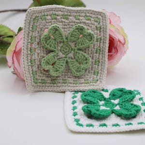 May include: Two crocheted square coasters. One is off-white with a green four-leaf clover design in the center. The other is white with a green clover and border. A pink rose and green leaves are in the background.