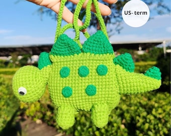 May include: A handmade, green crochet dinosaur-shaped bag with a strap. The bag features darker green accents on its back and head, with lighter green spots. The text "US-term" is visible on a white circle.