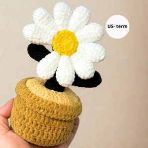 May include: A handmade crochet daisy flower with white petals and a yellow center, black stem, and a yellow-brown pot. The flower is a whimsical, decorative item.