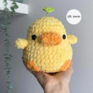 May include: A handmade, plush yellow duckling with orange accents on its beak and feet. The duckling has small black eyes and a green leaf sprouting from its head. The soft, textured material gives it a cuddly appearance.