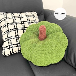 May include: A green, plush pumpkin-shaped cushion with a brown stem sits beside a cream cushion with a black plaid pattern. The cushions are on a black leather sofa.