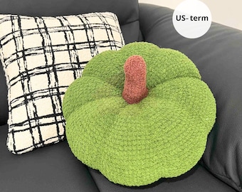 May include: A green, plush pumpkin-shaped cushion with a brown stem sits beside a cream cushion with a black plaid pattern. The cushions are on a black leather sofa.