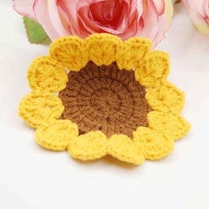 May include: A handmade crochet sunflower coaster with a brown center and yellow petals. The coaster is round and has a textured appearance. The sunflower is a decorative item, perfect for adding a touch of nature to any home decor.