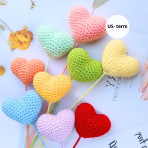 May include: A collection of colorful, crocheted heart-shaped decorations on sticks. The hearts are in various colors, including pink, green, yellow, orange, blue, and red. The image includes the text "US-term".