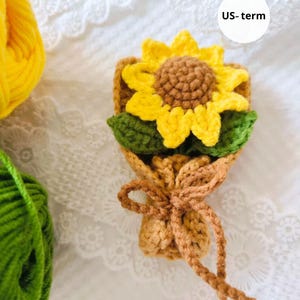 May include: A handmade crochet sunflower bouquet. The bouquet features a bright yellow sunflower with a brown center, green leaves, and a tan wrapping tied with a bow. The text "US-term" is in a white circle.