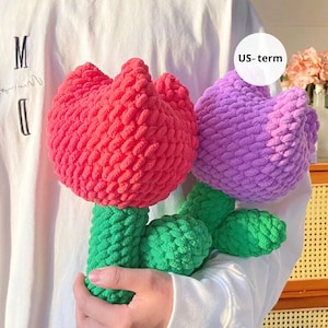 May include: Two handmade crochet flowers. One is a vibrant red, the other a soft purple. Both have green stems and leaves. The flowers are made with a thick, textured yarn, giving them a plush appearance. The background is a plain white shirt.