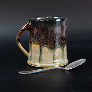 May include: A ceramic mug with a dark brown and black glaze featuring a drip pattern that reveals a lighter tan base color. The mug has a curved handle and a small circular emblem near the rim. A silver-colored metal spoon lies in front.