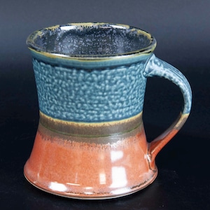 May include: A handmade ceramic mug with a textured blue upper section, transitioning to a brown and orange lower section. The mug has a sturdy handle and a dark blue rim. The mug is approximately 4 inches tall.