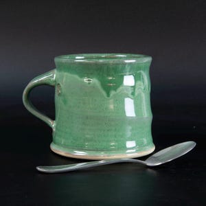 May include: A ceramic mug with a glossy, light green glaze featuring subtle darker green speckles and a smooth, rounded handle. The mug has a slightly flared rim and a thick base. A polished silver-colored metal spoon rests horizontally in front.