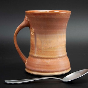 May include: A handmade, brown ceramic mug with a large handle and a wide base. The mug has a textured surface and a gradient color scheme, transitioning from a darker brown at the top to a lighter tan at the bottom. A silver spoon rests beside the mug.