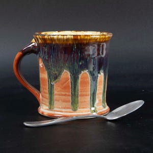 May include: A ceramic mug with a handle, featuring a terracotta-colored base and a dark blue and brown glaze that drips down the sides. A silver-colored metal spoon is positioned horizontally in front of the mug on a dark surface.