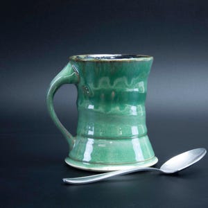 May include: A green ceramic mug with a unique, slightly asymmetrical handle. The mug has a glazed finish and a slightly flared base. A silver spoon is placed next to the mug.