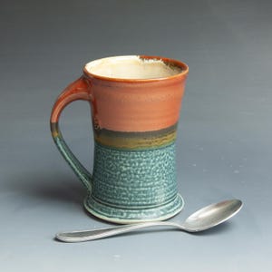 May include: A handmade ceramic mug with a textured teal base, a brown middle section, and a reddish-orange top. The handle is teal and brown. A silver spoon rests beside the mug. The mug is designed for beverages.