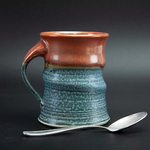 May include: Handmade ceramic mug with a textured blue body and a reddish-brown rim. The mug features a curved handle and a small, decorative swirl. A silver spoon rests beside the mug, suggesting its use for beverages.